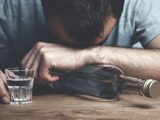 Mężczyzna z głową opartą o butelkę, obok kieliszek z alkoholem. Wygląda na to, że doświadcza zatrucia alkoholowego.