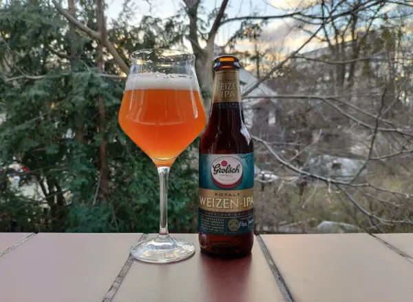 Szklanka z bursztynowym piwem Grolsch Royale Weizen IPA i butelka obok, na tle drzew i nieba.